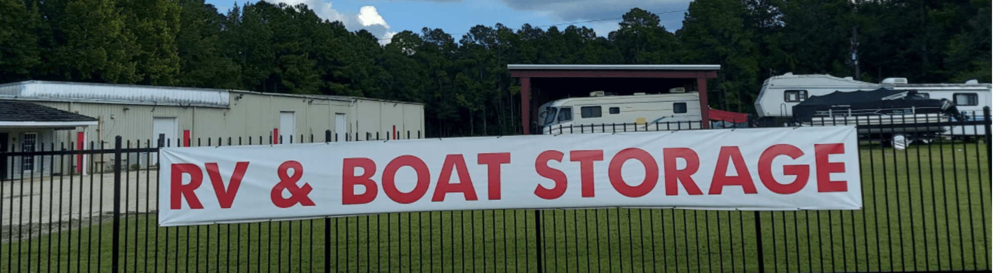 Motorhome, fifth-wheel camper, and pontoon boat parked behind the RV & Boat Storage fence banner